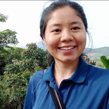 Young asian woman is wearing a blue collared shirt with her hair pulled back. She is standing in front of trees in the refugee camp she currently resides. 